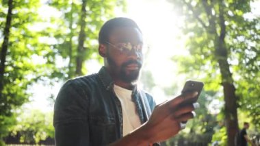 Ciddi afro-amerikan genç çekici adam telefon kullanıyor güneş ışığı altında duruyor günbatımı cep telefonu bilgisayar moda interneti teknoloji işadamları siyah şehir gezgini