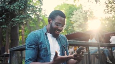 Mutlu Afro-Amerikan genç çekici adam, telefon kullanıyor, gün batımında caddede duruyor, cep telefonu, bilgisayar, internet, park teknolojisi, iş adamları, siyahi şehir gezgini.