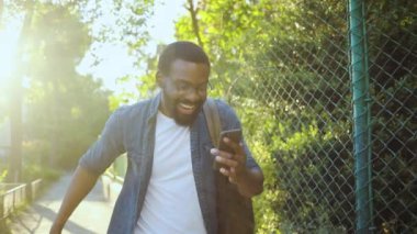 Gözlüklü, mutlu, Afro-Amerikan genç, gözlüklü, sokakta yürüyen, gülümseyen, güneş ışığı sıçrayan, günbatımı, cep telefonu moda interneti teknoloji işadamları, siyahi, genç şehir gezginleri.