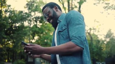 Afro-Amerikan genç çekici erkek gözlüklerini kapat. Sokakta yürü, gülümse, gün batımı, cep telefonu moda interneti. Teknoloji işadamları, siyahi şehir gezgini.