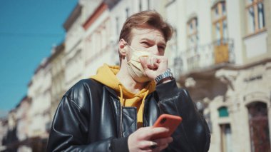 Sarışın, şık, beyaz, tıbbi maske takan, akıllı telefon kullanan, yüzünü kötü bir şekilde öksüren açık havada koronavirüs belirtileri gösteren bir adam. Salgın. Sağlık hizmetleri.