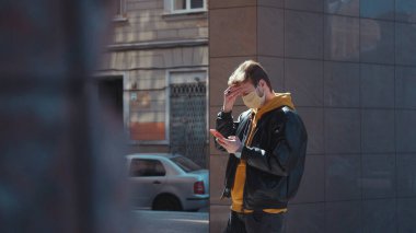 Şehir merkezinde akıllı telefon kullanan yüz maskeli genç bir adam. Sağlıksız üzgün adam kötü baş ağrısı çekiyor. Sağlık hizmetleri. Coronavirüs.