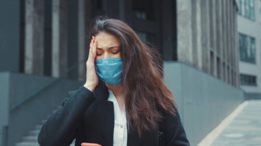 Sağlıksız, üzgün, beyaz, tıbbi maske takan ve baş ağrısı çeken genç bir kadının portresi sokakta şakaklarına dokunuyor. Karantina.