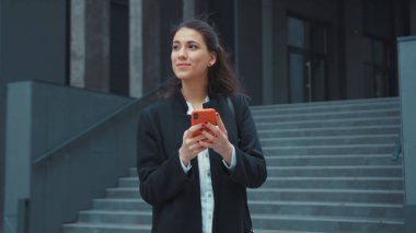 Binanın dışında akıllı telefon uygulaması kullanarak yürüyen genç bir bayanın sosyal medyada neşeli ve rahat bir şekilde mesajlaşması..