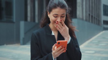 Kafası karışmış beyaz bir kadın akıllı telefondan haber zaman çizelgesi okuyor. Şaşırmış bir yüz ifadesiyle ofis binasının önünde durmakta tereddüt ediyor..