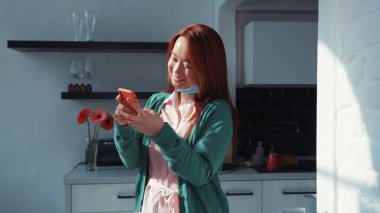 Neşeli Çinli, genç, kızıl saçlı, sıradan kıyafetler giyen, sosyal medyada akıllı telefon ağı kuran, mutfakta duran güzel bir kadın. Sosyal mesafe. Evde kal..