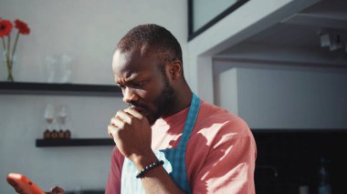 Üzüntülü üzgün siyah adam cep telefonundan olumsuz haberler okuyor, hayal kırıklığına uğruyor ve sorun hakkında kötü hissediyor. Mutfaktaki adam. Şok içeriği, kötü haber.