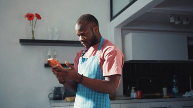 Aperatif giymiş Afro-Amerikan bir adam akıllı telefondan zaman çizelgesi okuyor ve kötü haber içeriğiyle hayal kırıklığına uğruyor. Mutfaktaki adam. Hayal kırıklığı. Teknoloji.