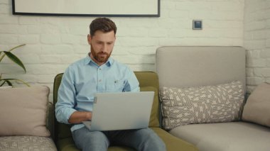 Çekici, mutlu adam evdeki modern oturma odasındaki koltukta oturmuş bilgisayarlı sohbet gülümsüyor yavaş çekimde işi bitiriyor.