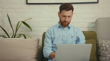 Çekici, mutlu adam evdeki modern oturma odasındaki koltukta oturmuş bilgisayarlı sohbet gülümsüyor yavaş çekimde işi bitiriyor.