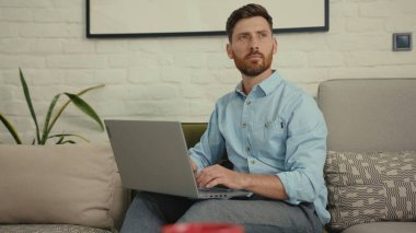 Bilgisayarda daktilo kullanan yakışıklı adam evdeki modern oturma odasındaki koltukta oturuyor bilgisayarlı sohbet gülüşü yavaş çekim kapat.