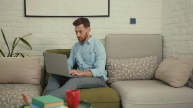 Bilgisayarda daktilo kullanan yakışıklı adam evdeki modern oturma odasındaki koltukta oturuyor bilgisayarlı sohbet gülüşü yavaş çekim kapat.