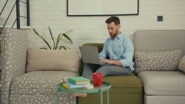 Bilgisayarda yazı yazan mutlu adam elini sallayan kameraya bak. Ev bilgisayarının modern oturma odasındaki koltukta oturan bir şeyi göstermek istiyor. Yavaş çekimde kapat.