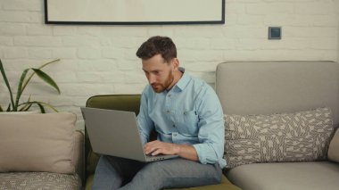 Bilgisayarda yazı yazan mutlu adam elini sallayan kameraya bak. Ev bilgisayarının modern oturma odasındaki koltukta oturan bir şeyi göstermek istiyor. Yavaş çekimde kapat.