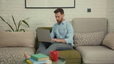 Bilgisayarda yazı yazan mutlu adam elini sallayan kameraya bak. Ev bilgisayarının modern oturma odasındaki koltukta oturan bir şeyi göstermek istiyor. Yavaş çekimde kapat.