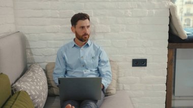 Bilgisayarda daktilo kullanan yakışıklı adam evdeki modern oturma odasındaki kanepede uzanıyor bilgisayarlı sohbet gülüşü yavaş çekim kapat.