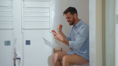Mutlu iş adamı tuvalette oturup gülümseyen cep telefonu akıllı telefon internet ev tuvaleti yavaş çekim banyoyu kapatıyor.