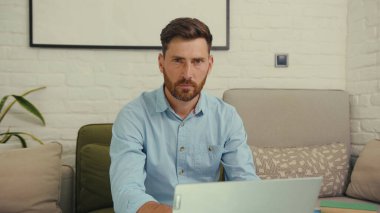 Portre çekici bir adam kameraya bakıyor ciddi işçi dizüstü bilgisayarı koltukta oturuyor internet girişimcisi bilgisayarı yavaş çekimde kapat