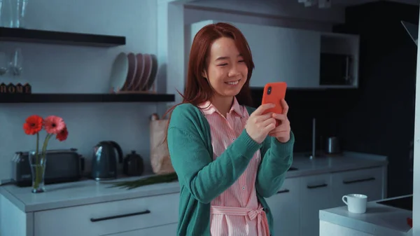 Attractive cute Asian young woman in mask stands in the kitchen messaging on smartphone social media application. Stay at home. Social distance. Communication concept.