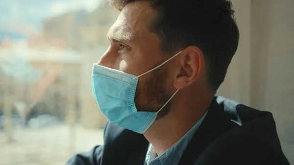 Sad businessman remove medical mask at home sitting on windowsill and looking at street on quarantine COVID-19 pandemic epidemic depressed portrait slow motion