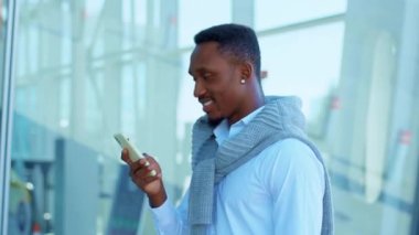 Afro-Amerikalı adam havaalanının yakınında yürürken akıllı telefon kullanıyor. Etrafına bakıp gülümsüyor. Kendine güvenen genç bir lider portresi. Açık havada. Yavaş çekim