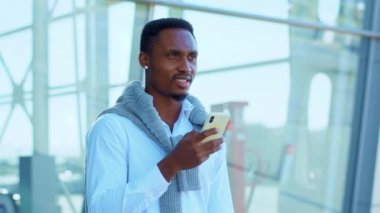 Kapatın, Afro-Amerikalı adam havaalanının yakınında yürürken akıllı telefon kullanın. Etrafınıza bakıp gülümseyin. Kendine güvenen genç bir lider portresi. Açık havada. Yavaş çekim