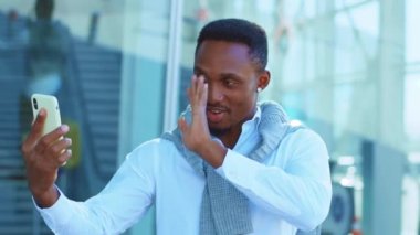 Mutlu Afro-Amerikan adam video konuşma için akıllı telefonu kullanıyor. Havaalanı yakınlarında gülümsüyor. Kendine güvenen genç bir lider portresi. Açık havada. Yavaş çekim