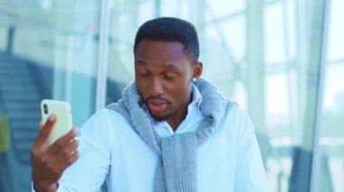 Sinirli Afrikalı Amerikalı adam, havaalanının yakınındaki telefon kulübesinde görüntülü konuşma için akıllı telefon kullanıyor. Kendine güvenen genç bir lider portresi. Açık havada. Yavaş çekim