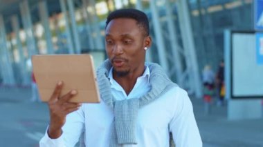 Yakışıklı Afro-Amerikalı gülümseyen genç bir portre. Tablet bilgisayar kullanıyor. Açık havada, havaalanının yakınında mutlu hissediyor. Dijital tablet ağ bilgisayar yazılımı. Kapatın. Yavaş çekim