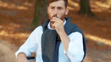 Yüzünü kapat. Tekerlekli sandalyedeki üzgün, çekici genç adam güneş ışığında güzel bir parkta sigara içiyor. Sakallı yakışıklı engelli yürüyüşü. Portre. Yavaş çekim