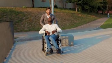Mutlu Afro-Amerikan erkeği arkadaşıyla sonbahar parkında tekerlekli sandalyede iyi eğlenceler. Yürüyen çekici sağlık araçlarını birlikte destekleyin. Yavaş çekim