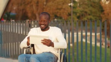 Tekerlekli sandalyedeki gülümseyen Afro-Amerikan adam güneş ışığı engelli tıbbının parkında elinde tablet bilgisayarla. Muhtemel özürlü engelli interneti. Yavaş çekim