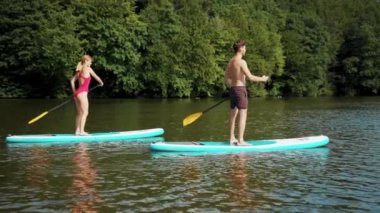 Yandan çekilen adam ve kadın mayo giyer ve gölde yüzer. Kürek çeken spor sörfçüleri için kürek çeker.