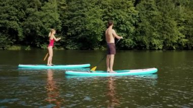 Genç adam ve kadın mayo giymiş, yemek masasında duruyor ve yüzme sporu sörfçüleri için kürek çeken arkadaşlarıyla gölde yüzüyor.