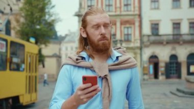 Sakallı hippi sıradan iş adamı güzel şehir merkezinde akıllı telefon kullanarak yürüyor. Arka planda mimari cazibesi. Tramvay hareket ediyor. Şehir manzarası. İnsanlar.
