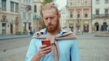 Şok olmuş bir ifadeyle kameraya bakan bir adamın akıllı telefondan mesaj almasına şaşırmış. Şehir manzarası. Açık havada. Yanlış anlaşılma.