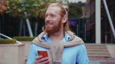 Milenyum zencefili çekici adam sokaklarda cep telefonu iletişimi uygulamasını kullanarak gülümsüyor ve şehirde kahve molası veriyor. İş Portresi.