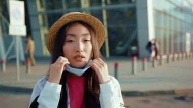 Havaalanı yakınlarındaki güzel Asyalı bayan kameraya bakarak yüz maskesi çıkarıyor. Sokaktaki çok ırklı, çarpıcı Çinli kadının açık hava portresi. Sağlık Hizmetleri.