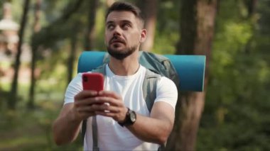 Ormanda sakallı ve sırt çantalı bir adam, ekranda bağlantı sinyali aramak için telefon kullanıyor. GPS başvurusu, turizm gezisi, kayıp güneş iletişimi, yavaş çekim telefon.