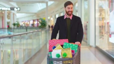 Mutlu genç çekici adam Noel hediyeleriyle yürüyor. Kutularla, yakışıklı gülücüklerle alışveriş yapıyor. Etrafına bak. Yeni yılı yavaş çekimde bitir.