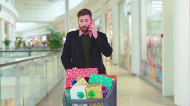 Noel hediyesi kutularıyla gergin sakallı bir adam alışveriş merkezinde telefonla konuşuyor. Noel ağacı alışverişi yapıyor, portre portresi yavaş çekimde.