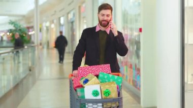 Noel hediyesi kutularıyla mutlu sakallı adam alışveriş merkezinde telefonla konuşuyor. Noel ağacı alışverişi yapıyor. Yüzü mobil hücreyi yavaş çekimde kapatıyor.