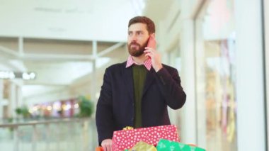 Noel hediyesi kutularıyla sakallı adam alışveriş merkezinde telefonla konuşuyor. Noel ağacı alışverişi yapıyor. Cep telefonunun yavaş çekimde mutlu bir portresi var.