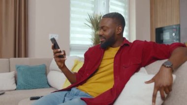 Kendine güveni olan çekici siyah bir adam, akıllı telefon online iletişim cihazı uygulaması kullanarak kanepede dinleniyor ve internette geziniyor. Modern daire. Cep telefonu olan insanlar.