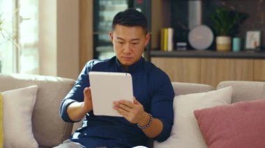 Çekici Asyalı genç adam evdeki kanepede oturan tablet bilgisayar kullanıyor. Çinli iş adamı çok yakışıklı. İletişim. Kapatın. Yavaş çekim