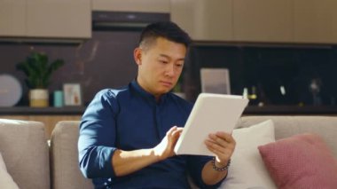 Evde ciddi Asyalı genç adam koltukta otururken tablet bilgisayar kullanıyor. Çinli iş adamı çok yakışıklı. İletişim. Kapatın. Yavaş çekim
