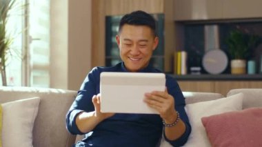 Gülümseyen Asyalı genç evdeki kanepede oturan tablet bilgisayar kullanıyor. Mutlu ol. Çinli iş adamı çok yakışıklı. İletişim. Kapatın. Yavaş çekim