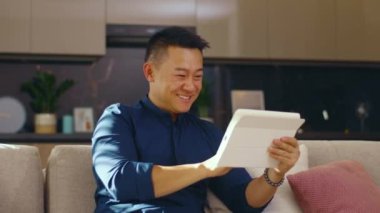 Mutlu Asyalı genç adam evdeki kanepede oturan tablet bilgisayarı kullanıyor. Çinli iş adamı çok yakışıklı. İletişim. Kapatın. Yavaş çekim