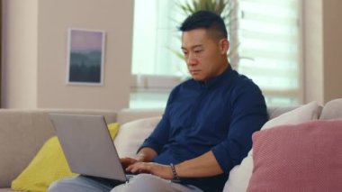 Kanepede oturan ciddi Asyalı adam evde dizüstü bilgisayar kullanıyor. Ağ işi görüntüleme elemanı iç teknoloji sosyal medya. Yavaş çekim