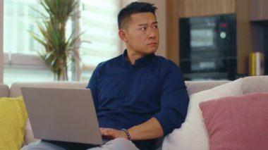 Kanepede oturan ciddi Asyalı adam evde dizüstü bilgisayar kullanıyor. Ağ işi görüntüleme elemanı iç teknoloji sosyal medya. Yavaş çekim